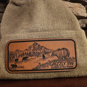 May include: A brown leather patch with a mountain scene and bears. The text "GRIZZLY 399 QUEEN of the Tetons" is on the patch.