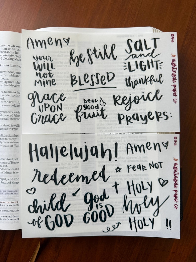 Pu&ograve; includere: Due fogli di adesivi bianchi con testo nero scritto a mano. Il foglio superiore include frasi come "Amen", "Be Still" e "Grace". Il foglio inferiore presenta "Hallelujah", "Redeemed" e "Child of God". Gli adesivi sono di "nightingale paper co."