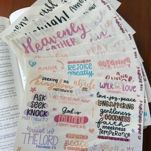 Pu&ograve; includere: Una pila di adesivi di carta traslucida con citazioni e frasi ispiratrici scritte a mano in vari colori. Gli adesivi includono frasi come "Heavenly", "God is Faithful" e "Perfect Peace".