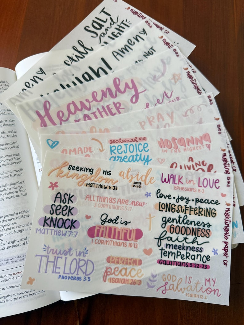 Pu&ograve; includere: Una pila di adesivi di carta traslucida con citazioni e frasi ispiratrici scritte a mano in vari colori. Gli adesivi includono frasi come "Heavenly", "God is Faithful" e "Perfect Peace".