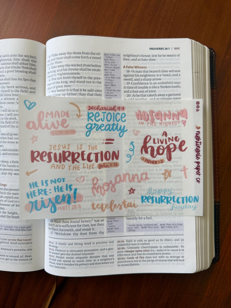 Pu&ograve; includere: Una Bibbia aperta su Proverbi 26:1, con diversi adesivi colorati scritti a mano. Gli adesivi presentano frasi come "Made Alive", "Rejoice Greatly" e "Ges&ugrave; &egrave; la Risurrezione e la Vita". Gli adesivi sono di nightingale paper co.