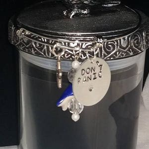 Può includere: Un barattolo di vetro nero per candele con un coperchio argentato che presenta una statuetta di un uccello. Il coperchio ha un design decorativo e un ciondolo con la scritta "Non farti prendere dal panico".