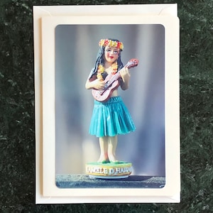May include: A figurine of a woman wearing a flower lei and a blue hula skirt, playing a ukulele. The figurine is standing on a base that says "Lanele o Hawaii".