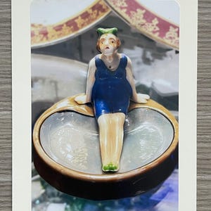 May include: A vintage ceramic figurine of a woman in a blue swimsuit sitting in a brown and white ceramic dish. The figurine has green hair and is wearing a green bow. The dish has a scalloped edge and a white interior.