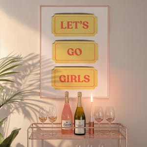 Könnte beinhalten: Gerahmter Druck mit den Worten „LET'S GO GIRLS“ in Rot auf gelben, ticketförmigen Hintergründen. Darunter ein Servierwagen mit Weinflaschen, Gläsern und einer brennenden Kerze. Der Wagen ist roségoldfarben.
