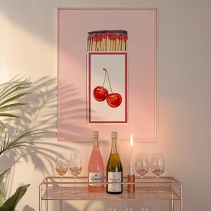 Könnte beinhalten: Ein rosa gerahmter Druck mit einer weißen Box, die eine Schachtel Streichhölzer und zwei rote Kirschen enthält. Zwei Flaschen Wein und zwei Weingläser stehen auf einem goldenen Barwagen mit einer Kerze in einem Glashalter.
