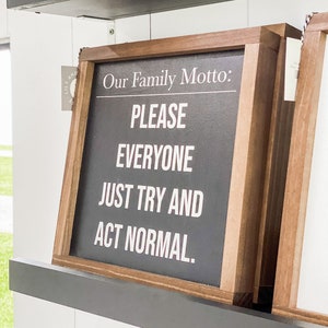 May include: A wooden framed sign with a black chalkboard insert. The sign reads "Our Family Motto: PLEASE EVERYONE JUST TRY AND ACT NORMAL."