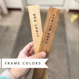 Pode incluir: Duas amostras de madeira, uma em carvalho claro e outra em nogueira escura, s&atilde;o seguradas na m&atilde;o. O texto "FRAME COLORS" &eacute; vis&iacute;vel abaixo das amostras.