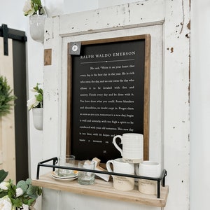 Pode incluir: Uma placa preta enquadrada com fundo branco e uma citaat de Ralph Waldo Emerson. A citaat diz: "Ele disse: 'Escreva isso em seu cora&ccedil;&atilde;o que cada dia &eacute; o melhor dia do ano. &Eacute; rico quem possui o dia, e ningu&eacute;m possui o dia que permite que seja invadido por preocupa&ccedil;&otilde;es e ansiedade. Termine cada dia e fa&ccedil;a as pazes com ele. Voc&ecirc; fez o que p&ocirc;de. Sem d&uacute;vida, alguns erros e absurdos se infiltraram. Esque&ccedil;a-os o mais r&aacute;pido poss&iacute;vel, amanh&atilde; &eacute; um novo dia; comece bem e com serenidade, com um esp&iacute;rito muito elevado para ser sobrecarregado por suas velhas bobagens. Este novo dia &eacute; muito precioso, com suas esperan&ccedil;as e...'" A placa est&aacute; pendurada em uma porta branca com uma prateleira embaixo. A prateleira &eacute; de madeira e tem uma moldura de metal preto. H&aacute; tr&ecirc;s canecas de cer&acirc;mica branca na prateleira.