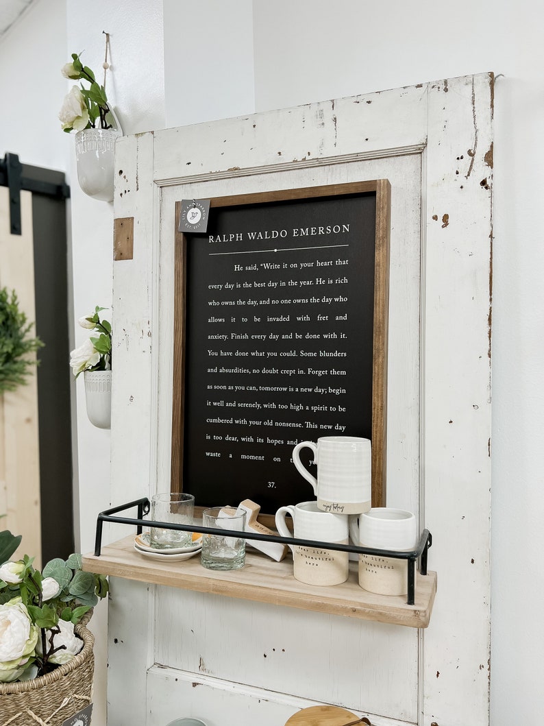 Pode incluir: Uma placa preta enquadrada com fundo branco e uma citaat de Ralph Waldo Emerson. A citaat diz: "Ele disse: 'Escreva isso em seu cora&ccedil;&atilde;o que cada dia &eacute; o melhor dia do ano. &Eacute; rico quem possui o dia, e ningu&eacute;m possui o dia que permite que seja invadido por preocupa&ccedil;&otilde;es e ansiedade. Termine cada dia e fa&ccedil;a as pazes com ele. Voc&ecirc; fez o que p&ocirc;de. Sem d&uacute;vida, alguns erros e absurdos se infiltraram. Esque&ccedil;a-os o mais r&aacute;pido poss&iacute;vel, amanh&atilde; &eacute; um novo dia; comece bem e com serenidade, com um esp&iacute;rito muito elevado para ser sobrecarregado por suas velhas bobagens. Este novo dia &eacute; muito precioso, com suas esperan&ccedil;as e...'" A placa est&aacute; pendurada em uma porta branca com uma prateleira embaixo. A prateleira &eacute; de madeira e tem uma moldura de metal preto. H&aacute; tr&ecirc;s canecas de cer&acirc;mica branca na prateleira.
