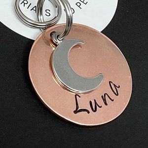 May include: Copper pet tag with a silver crescent moon charm and the name "Luna" engraved on it. The tag is on a black background with a white circle that says "Boonsepia Pet Tags".