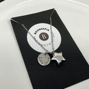 Pu&ograve; includere: Una collana argentata con un ciondolo a forma di stella e un ciondolo rotondo con inciso "Lola Bean". La collana &egrave; esposta su un cartoncino nero con un logo circolare bianco che recita "BOONSEPIA MEMORIALS / ID TAGS".