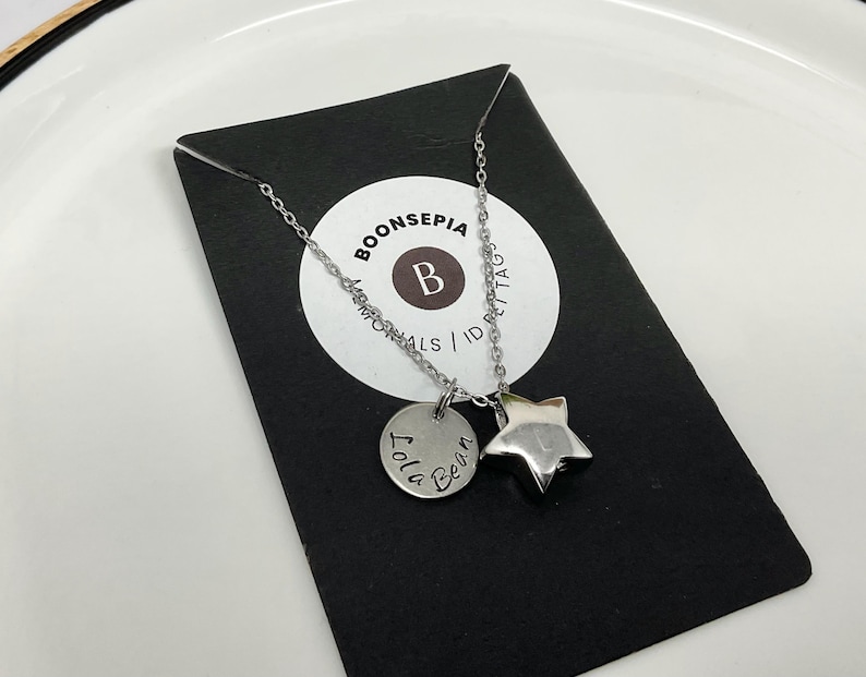 Pu&ograve; includere: Una collana argentata con un ciondolo a forma di stella e un ciondolo rotondo con inciso "Lola Bean". La collana &egrave; esposta su un cartoncino nero con un logo circolare bianco che recita "BOONSEPIA MEMORIALS / ID TAGS".