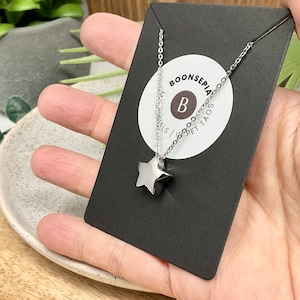 Pu&ograve; includere: Una collana con pendente a stella argentata su una carta nera. La collana ha una catena sottile e un pendente a forma di stella a cinque punte. La carta ha un logo circolare bianco con la lettera "B" e il testo "BOONSEPIA MATERIALS | PET TAGS".