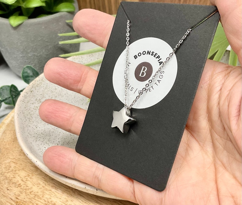 Pu&ograve; includere: Una collana con pendente a stella argentata su una carta nera. La collana ha una catena sottile e un pendente a forma di stella a cinque punte. La carta ha un logo circolare bianco con la lettera "B" e il testo "BOONSEPIA MATERIALS | PET TAGS".