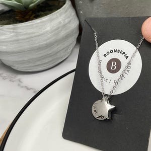 Pu&ograve; includere: Una collana color argento con un ciondolo a forma di stella e un piccolo tag rotondo. Il tag ha inciso il numero "10+". La collana &egrave; esposta su una carta nera con il marchio "BOONSEPIA" e altro testo.