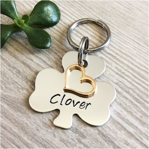 May include: Silver clover-shaped pet tag with a gold heart charm and the name "Clover" engraved on it. The tag is attached to a silver key ring.