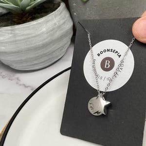 Pu&ograve; includere: Una collana color argento con un ciondolo a forma di stella e un piccolo ciondolo rotondo. Il ciondolo ha il testo "LOL" e la collana &egrave; esposta su una carta nera con il testo "BOONSEPIA MEMORIALS / ID & PET TAGS".
