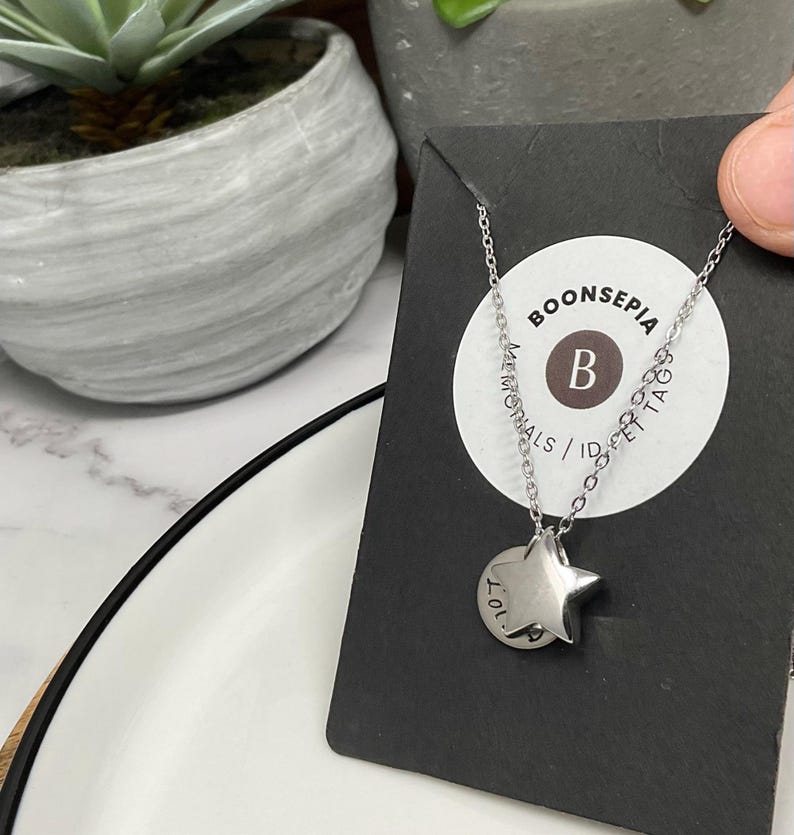 Pu&ograve; includere: Una collana color argento con un ciondolo a forma di stella e un piccolo ciondolo rotondo. Il ciondolo ha il testo "LOL" e la collana &egrave; esposta su una carta nera con il testo "BOONSEPIA MEMORIALS / ID & PET TAGS".