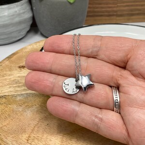 Pu&ograve; includere: Una collana d'argento con un ciondolo a forma di stella e un ciondolo rotondo. Il ciondolo rotondo ha il testo "Jaci Le". La collana &egrave; su una superficie di legno.