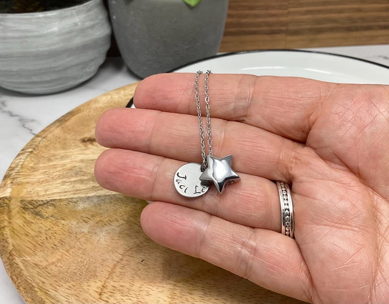 Pu&ograve; includere: Una collana d'argento con un ciondolo a forma di stella e un ciondolo rotondo. Il ciondolo rotondo ha il testo "Jaci Le". La collana &egrave; su una superficie di legno.
