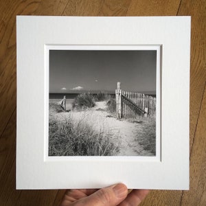 Op de afbeelding: Zwart-witfoto van een strandscène. Een zanderig pad leidt door hoog gras en een houten hek naar de oceaan. De lucht is helder met een paar wolken. De foto is wit ingelijst.