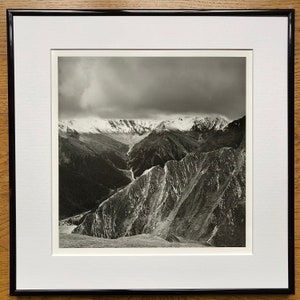 May include: A black and white photograph of a mountain range with snow-capped peaks and a valley in the foreground. The sky is cloudy and the mountains are rugged and dramatic.