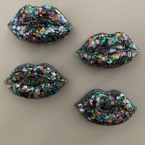 May include: Four black resin lips with a clear, glittery, iridescent finish. The glitter is in shades of pink, green, blue, gold, and black.
