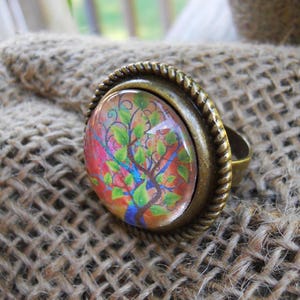 May include: An antique brass ring with a large, oval-shaped, colorful tree design under a glass dome. The ring has a braided rope-like border around the tree image. The ring is set on a burlap surface.