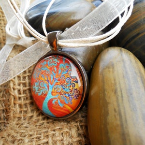 May include: A round pendant necklace with a colorful tree design. The pendant has a silver-colored metal frame and hangs on a white cord and ribbon. The tree design features orange, blue, and yellow colors. The necklace is displayed on a textured, brown surface with smooth stones.