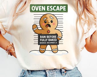 Könnte beinhalten: Cremefarbenes T-Shirt mit einem verwitterten Lebkuchenmann-Design. Die Grafik enthält den Text "OVEN ESCAPE" über dem Lebkuchenmann und "RAN BEFORE FULLY BAKED BOOKED: DECEMBER 25" darunter.