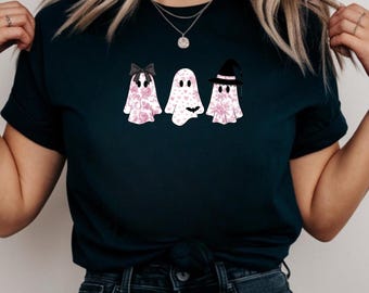 May include: A dark navy blue t-shirt with three ghost illustrations. The ghosts are white with pink floral patterns and black details, including a bow, a bat, and a witch's hat. The shirt is tied at the waist.