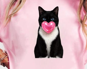 Puede incluir: Camiseta rosa con un gato blanco y negro de ojos azules sosteniendo un corazón rosa. El gato es el foco central del diseño. La camiseta es de color rosa liso.
