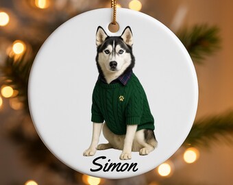 May include: A round, white ceramic ornament featuring a Siberian Husky wearing a green sweater with a paw print. The dog has black and white fur and blue eyes. The name "Simon" is printed in black script at the bottom. A gold string is attached for hanging.