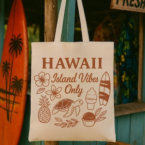 May include: Beige tote bag with brown text and illustrations. The text reads "HAWAII Island Vibes Only." Illustrations include a flower, pineapple, turtle, ice cream, and surfboard. The bag has white handles and is hanging outdoors.