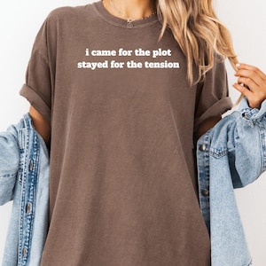 Może przedstawiać: Brązowy t-shirt z białym napisem "i came for the plot stayed for the tension". Koszulka jest połączona z dżinsową kurtką. Rękawy są podwinięte.