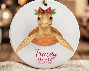 May include: A circular ceramic Christmas decoration. It depicts a sea turtle wearing a red and green tartan bow and greenery. The name "Tracey" and the year "2025" are written in red script below the turtle.