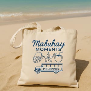 Può includere: Borsa tote color crema con testo e illustrazioni blu. Il testo recita "Mabuhay Moments" sopra un mango, una stella, una bevanda al cocco e una jeepney con la scritta "Philippines". Sfondo di spiaggia sabbiosa e oceano.