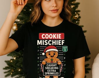 Könnte beinhalten: Schwarzes T-Shirt mit einem Lebkuchenmann-Motiv. Der Aufdruck lautet "COOKIE MISCHIEF" und "CAUGHT SNEAKING EXTRA SPRINKLES". Der Lebkuchenmann trägt einen Weihnachtsmannhut. Der Text "BOOKED: DECEMBER 25" ist ebenfalls enthalten.