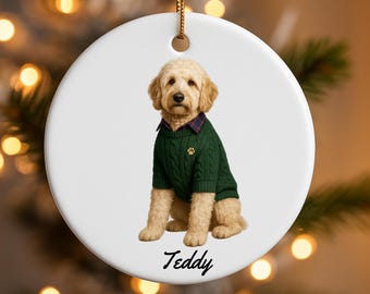 May include: A white ceramic ornament featuring a golden doodle dog wearing a green sweater with a paw print. The dog has a plaid collar. The name "Teddy" is written in script below the dog. The ornament hangs from a gold string.