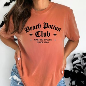 May include: A burnt orange t-shirt with the text "Beach Potion Club" in a gothic font, with the words "Casting Spells Since 1986" below. The shirt is worn with blue jeans.