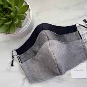 Puede incluir: Tres mascarillas, una negra, una gris oscuro y una gris claro, con el&aacute;sticos de oreja blancos y una etiqueta blanca que dice "Ten Choose Design".