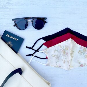 May include: A white tote bag with black handles, a passport with the word "PASSPORT" printed on the cover, a pair of tortoiseshell sunglasses, and three face masks. The face masks are layered on top of each other. The top mask is white with a floral pattern, the middle mask is red, and the bottom mask is black.