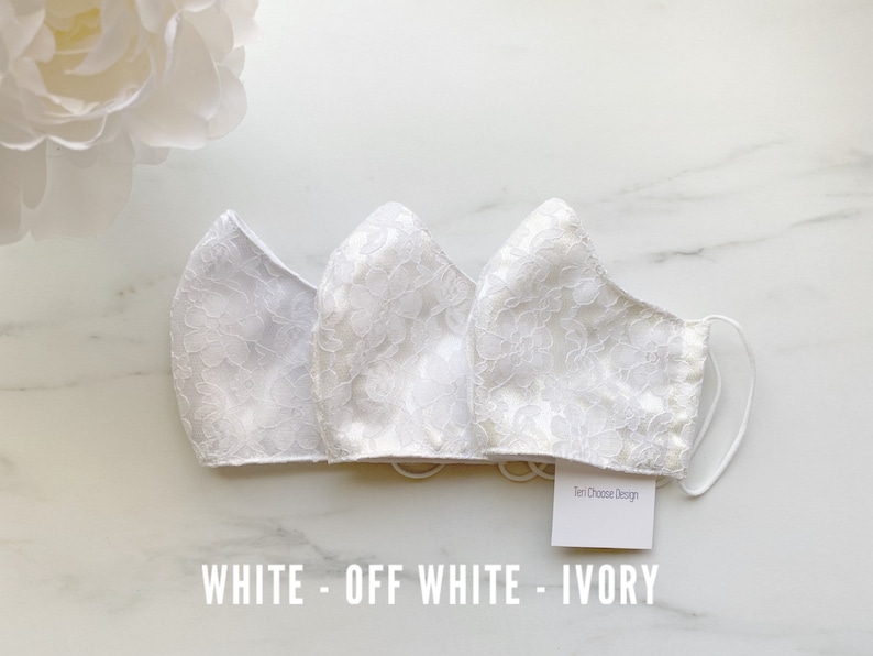 Puede incluir: Tres mascarillas de encaje blanco, una en blanco, otra en blanco roto y otra en marfil. Las mascarillas est&aacute;n dispuestas sobre una superficie de m&aacute;rmol blanco. Una peque&ntilde;a etiqueta blanca con el texto "Ten Choose Design" es visible en la esquina inferior derecha de la imagen. El texto "WHITE - OFF WHITE - IVORY" est&aacute; impreso debajo de las mascarillas.