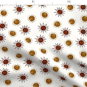 May include: A white fabric with a repeating pattern of brown and gold suns. The suns have a simple, hand-drawn style with radiating lines.