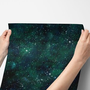 K&ouml;nnte beinhalten: Die H&auml;nde einer Person halten ein Blatt dunkelgr&uuml;nes und blaues Tapetenpapier mit einem Galaxie-Druck. Die Tapete ist mit wei&szlig;en Sternen bedeckt.