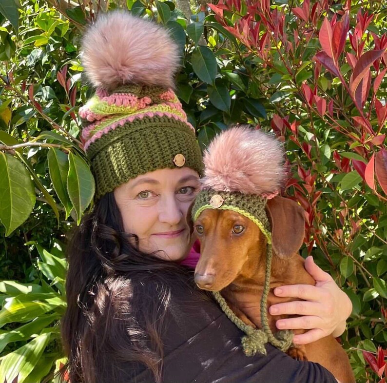 Puede incluir: Una mujer y un perro dachshund llevan gorros de punto verde, rosa y amarillo a juego con pompones. La mujer est&aacute; sosteniendo al perro en sus brazos.