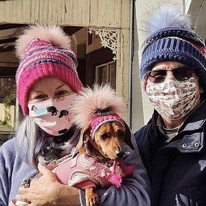 Puede incluir: Dos personas con gorros de invierno y mascarillas caminan con un peque&ntilde;o dachshund que lleva un gorro rosa y un jersey rosa. El dachshund est&aacute; siendo sostenido por la persona de la izquierda.
