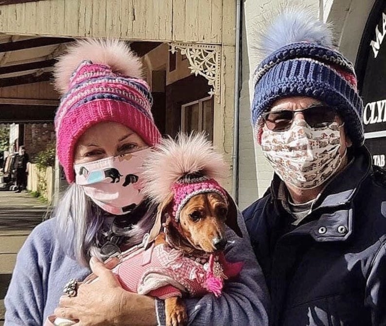 Puede incluir: Dos personas con gorros de invierno y mascarillas caminan con un peque&ntilde;o dachshund que lleva un gorro rosa y un jersey rosa. El dachshund est&aacute; siendo sostenido por la persona de la izquierda.