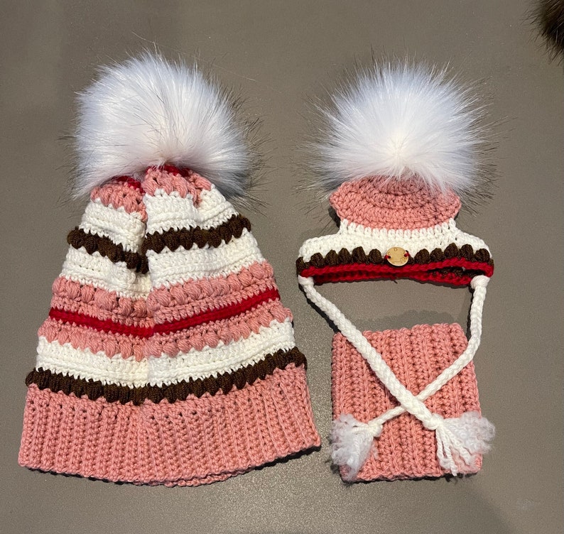 Puede incluir: Dos gorros de punto con pompones blancos. El gorro m&aacute;s grande es rosa, blanco, marr&oacute;n y rojo con un patr&oacute;n a rayas. El gorro m&aacute;s peque&ntilde;o es rosa, blanco, marr&oacute;n y rojo con un patr&oacute;n a rayas y un cuello de punto rosa a juego.